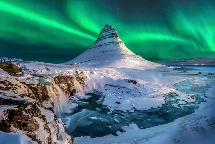 A breathtaking view of the Northern Lights illuminating the night sky over Kirkjufell mountain in Iceland, with a frozen waterfall and snow-covered landscape in the foreground, showcasing the stunning natural beauty of Iceland in March.