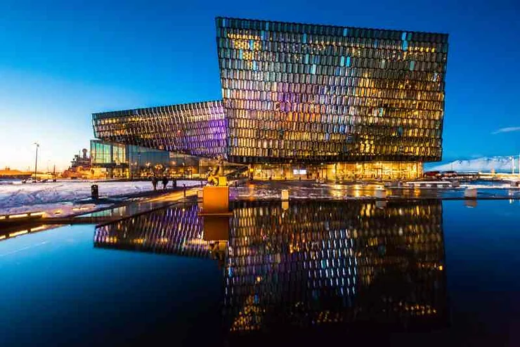 A stunning view of the Harpa Concert Hall in Reykjavik, Iceland, illuminated at night with colorful lights reflecting off its glass facade and the water in front, showcasing one of the popular activities to do in Iceland in March.