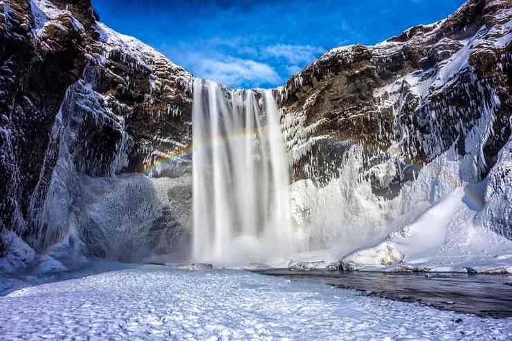 A majestic waterfall cascading down icy cliffs in Iceland in March. The falls are surrounded by snow and ice, creating a winter wonderland. A faint rainbow is visible in the mist, set against a bright blue sky.