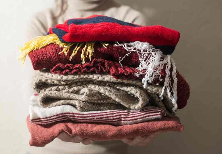 A person holding a stack of warm winter clothes, including sweaters, scarves, and woolen garments, ideal for the cold weather in Iceland in March.