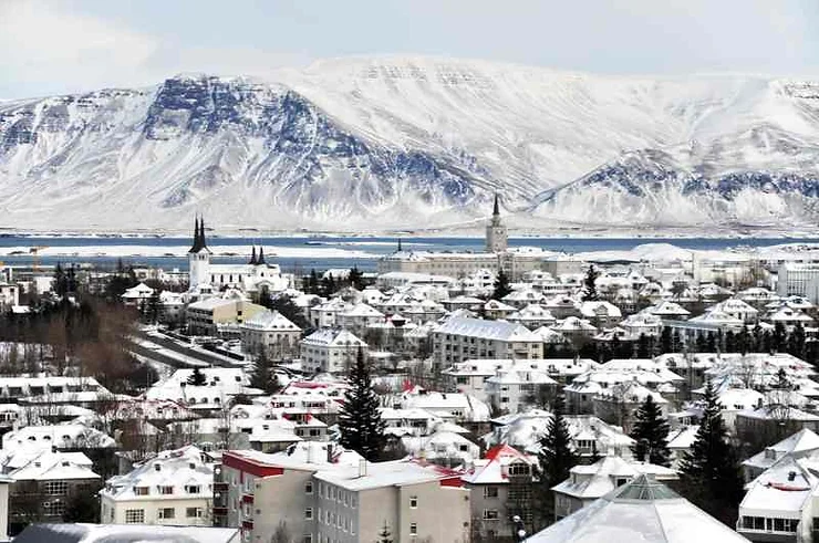 A panoramic view of Reykjavik, Iceland in March, covered in a blanket of snow. The city's rooftops and streets are coated with snow, with majestic snow-capped mountains and a clear sky in the background, showcasing the serene winter beauty of Iceland.