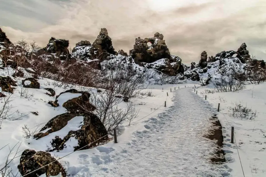 Snow-covered path leading through the unique volcanic rock formations of Dimmuborgir lava field in Iceland under a cloudy sky.