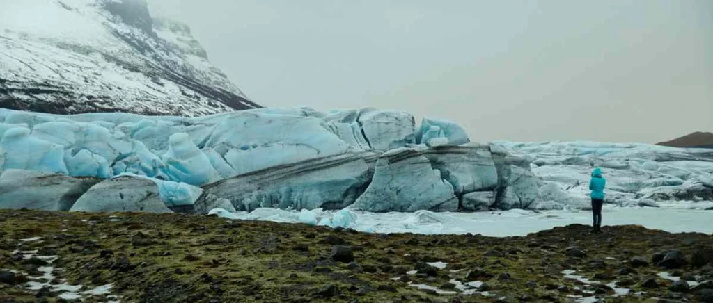 Person in blue jacket standing in front of a massive glacier in Iceland, with snow-covered mountains in the background.