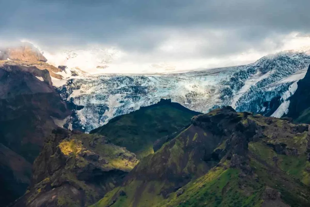 Lush green hills in the foreground with the majestic Mýrdalsjökull Glacier and snow-capped mountains in the background under a cloudy sky.