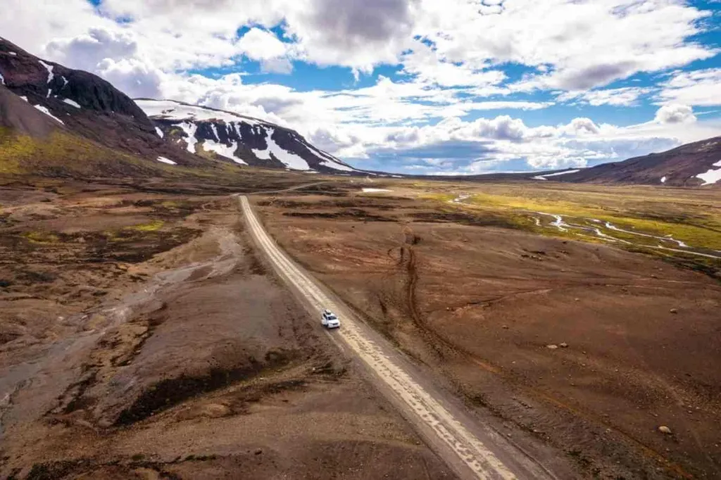 A white car driving on a dirt road through a barren, mountainous landscape in Iceland under a partly cloudy sky.