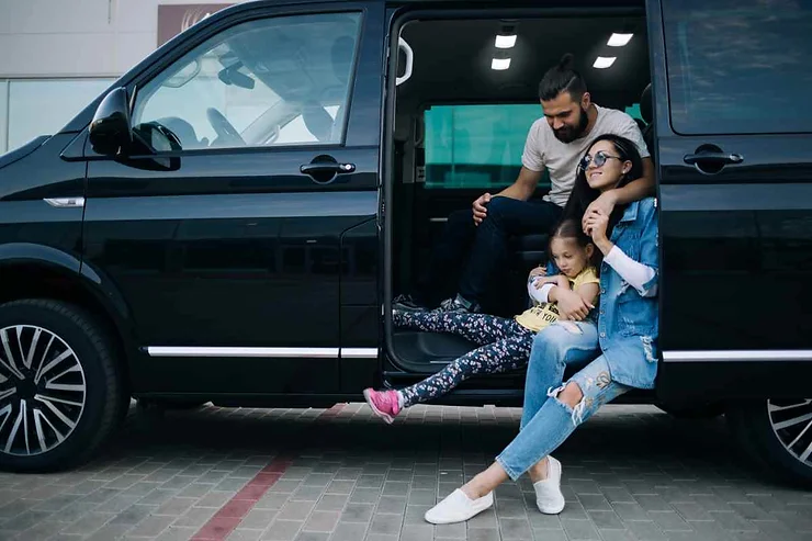 A family sitting by the open door of a black minivan. The father is sitting inside, while the mother and child are sitting on the edge, enjoying a moment together.