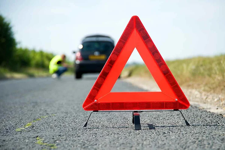 A red warning triangle placed on the road with a blurred view of a car and a person in a high-visibility jacket in the background.