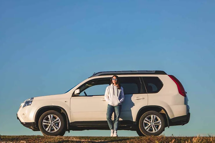 A person in a white hoodie and jeans standing next to a white SUV with a clear blue sky in the background.