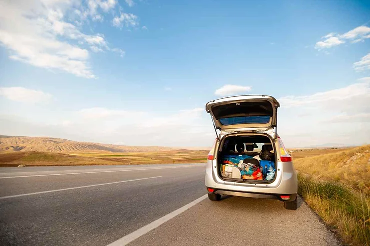 A silver SUV parked on the side of the road with its trunk open, showing packed luggage inside. The background features a wide open landscape under a blue sky with scattered clouds.