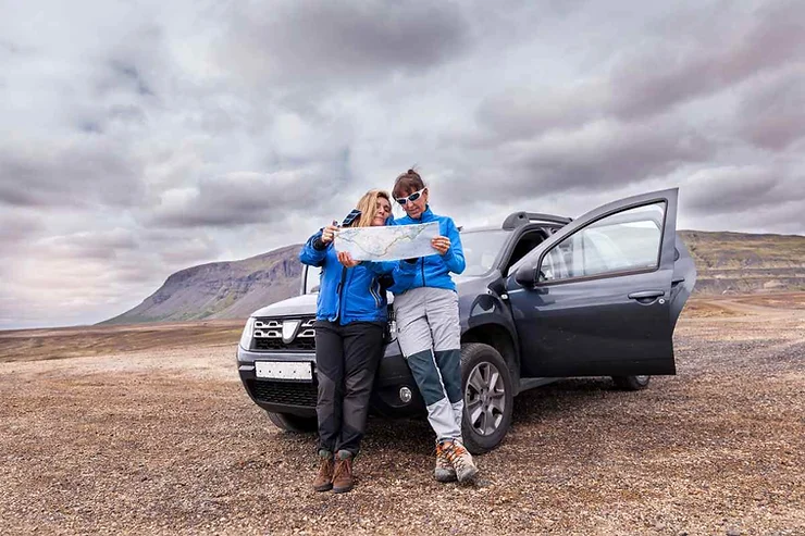 Two people wearing blue jackets standing next to a gray SUV on a barren landscape. They are looking at a map, planning their route. The SUV has its door open, and the background features a rugged mountainous terrain under a cloudy sky.