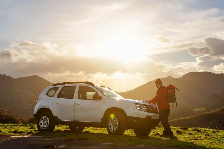 A person with a backpack standing next to a white SUV in a scenic outdoor setting with mountains and a bright sky in the background.