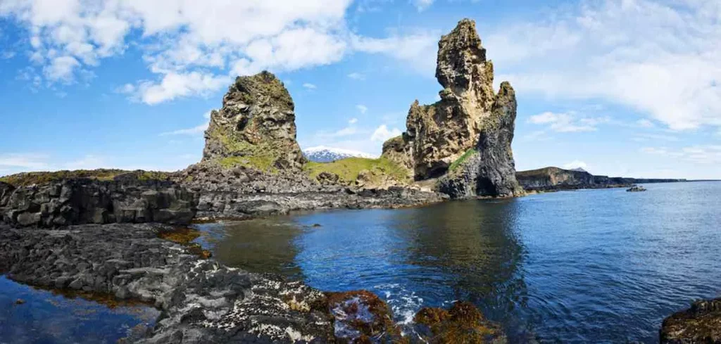 Dramatic view of Londrangar cliffs in Iceland with rugged coastline, dark volcanic rocks, and turbulent blue-green ocean waves under a cloudy sky.