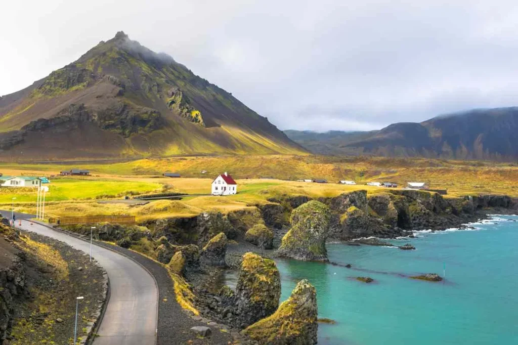 Beautiful coastal landscape in Iceland with a road leading to a white house, surrounded by green fields, rocky formations, and turquoise waters.