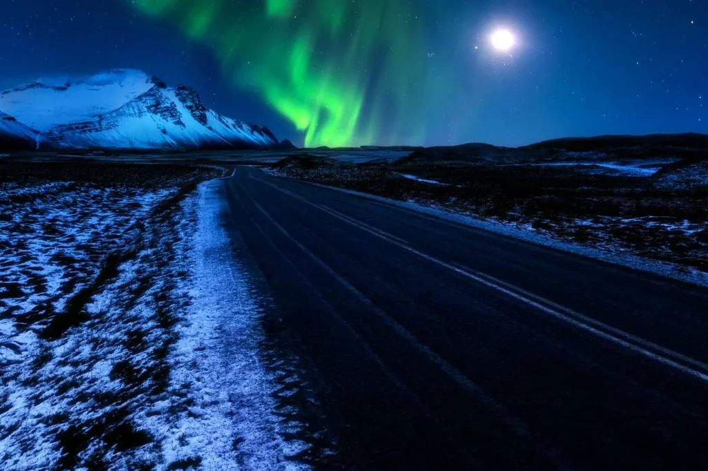 Northern Lights dancing over Iceland's Ring Road, with vibrant green auroras illuminating the night sky and the moon shining brightly.