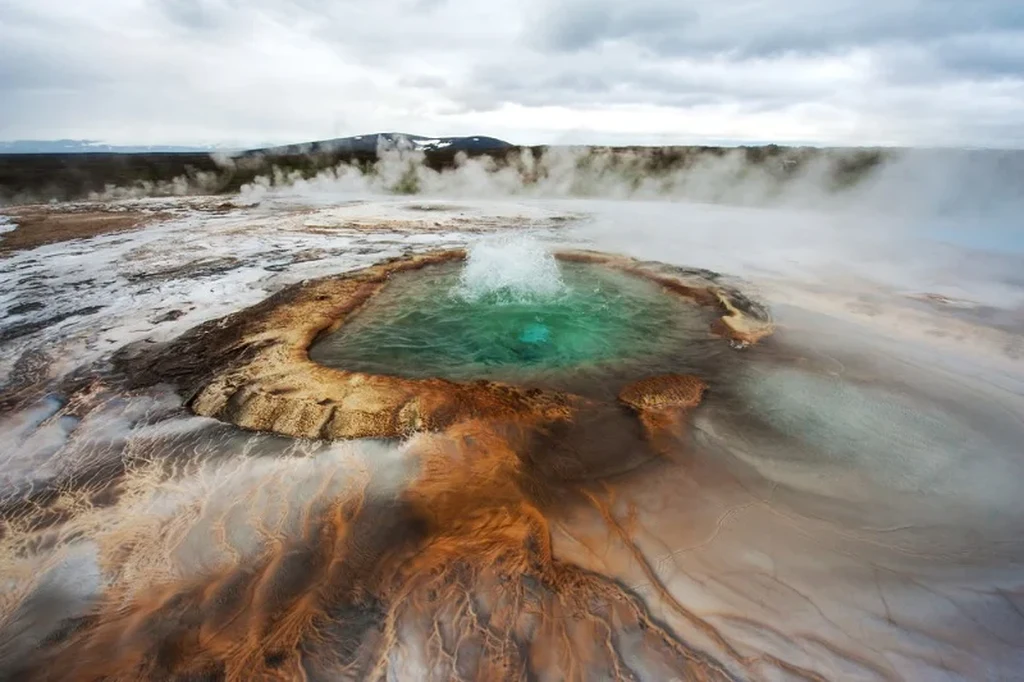 Geysir thermal area in Iceland, featuring a vibrant turquoise hot spring surrounded by steaming earth and dramatic mineral deposits.