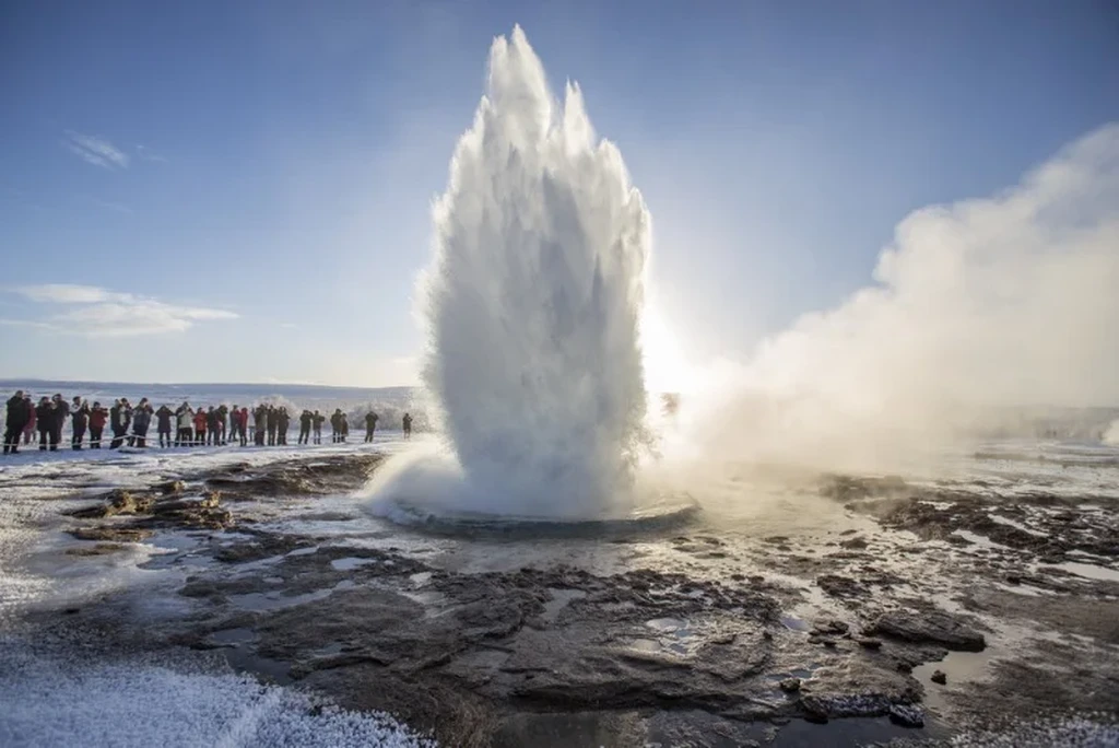 Strokkur geyser erupting in Iceland, surrounded by a crowd of tourists on a clear winter day, with steam rising into the blue sky.