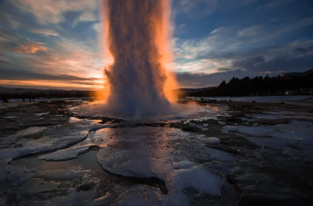 Strokkur geyser erupting at sunset in Iceland, creating a dramatic scene with water and steam illuminated by the golden light.