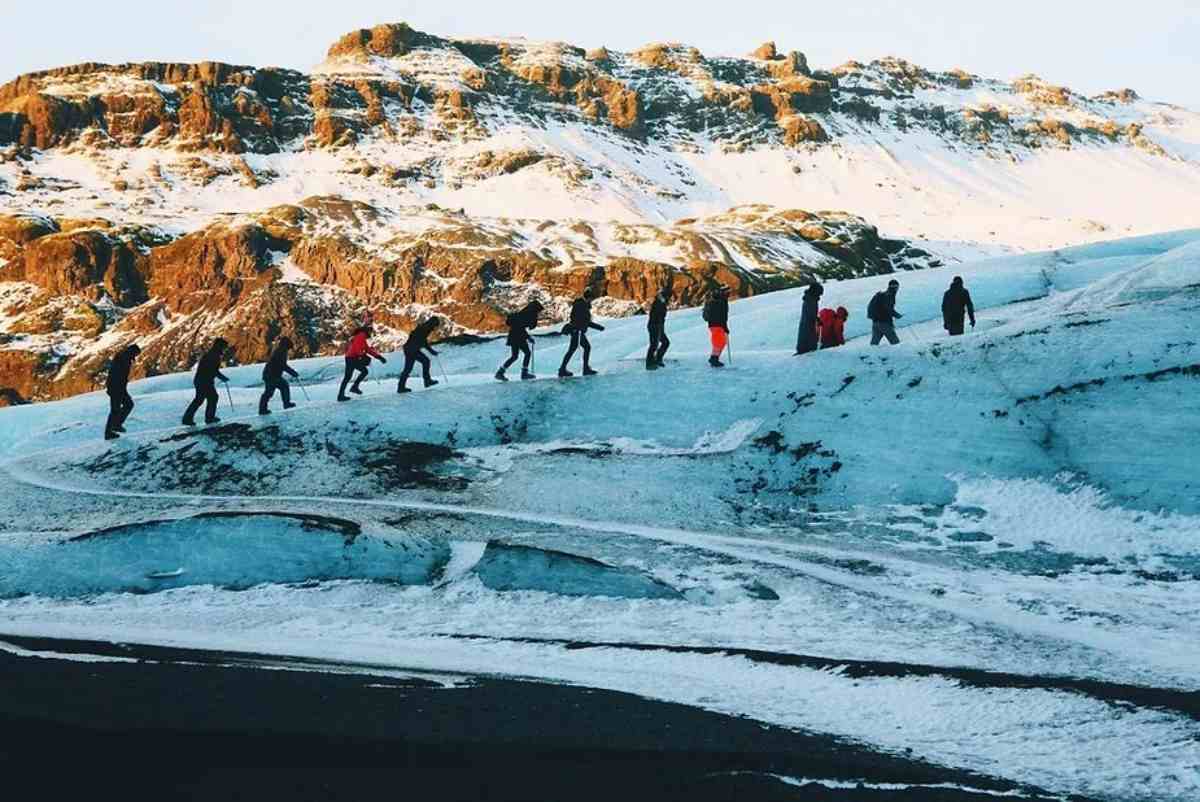 Group of tourists hiking on a glacier in Iceland, with rocky cliffs and snow-covered mountains in the background.