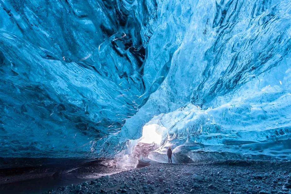 A person standing inside a stunning blue ice cave in Iceland, illuminated by natural light filtering through the ice, showcasing the beauty and scale of the icy formations.