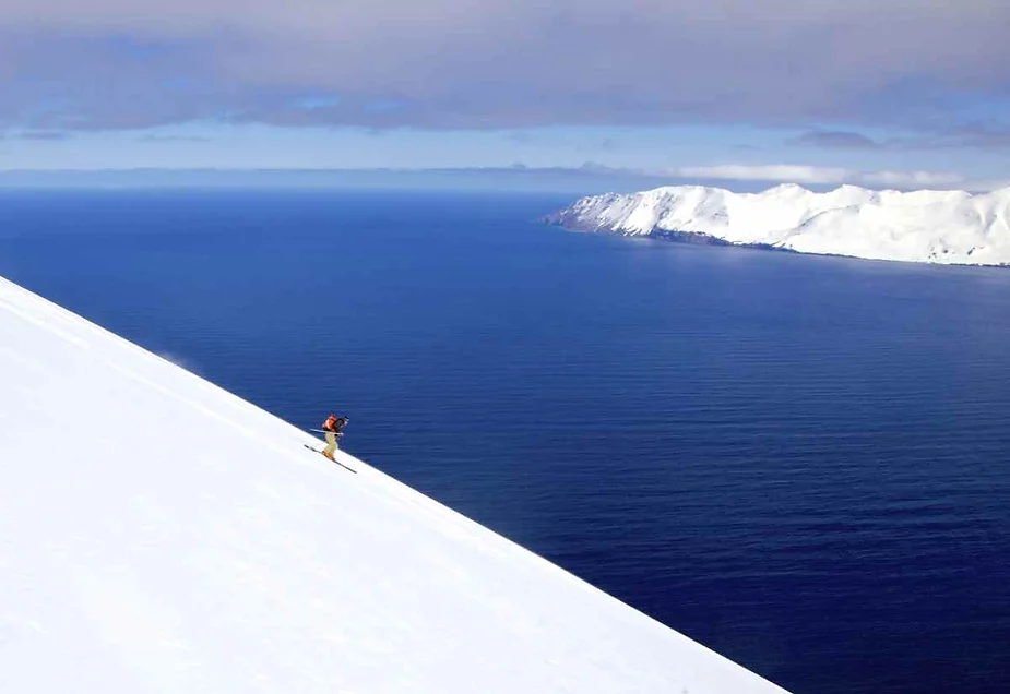 Lone skier descending a steep snowy slope with a breathtaking view of the blue ocean and snow-covered cliffs in Iceland.