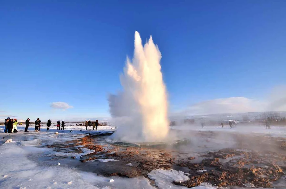 A dramatic scene of a geyser eruption in Iceland, surrounded by a group of mesmerized spectators against a clear blue sky.