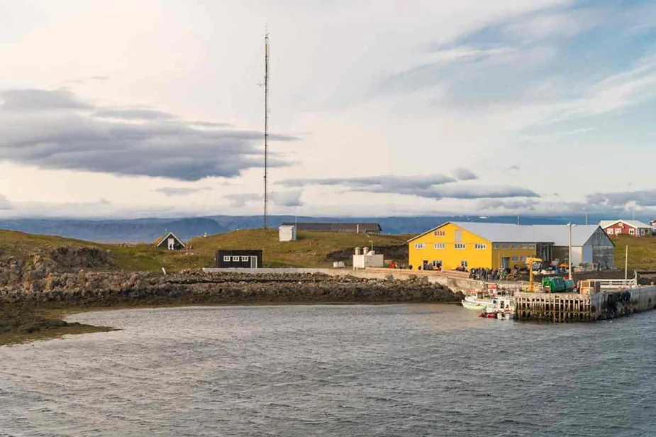 A serene coastal village in Iceland with a yellow building, a dock with boats, and a tall antenna against a backdrop of rolling hills and a cloudy sky.