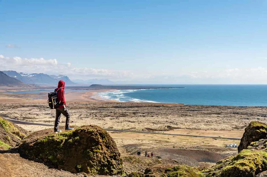 Hiker in a red jacket standing on a hill overlooking a stunning Icelandic coastline, with vast sandy beaches, rugged terrain, and clear blue skies in the background.