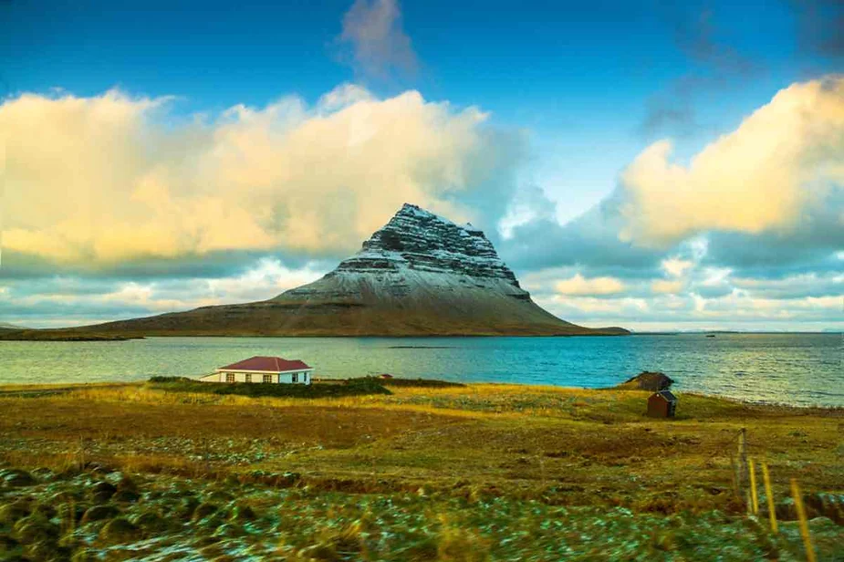 A picturesque scene in Iceland featuring a solitary house near a lake with the iconic Kirkjufell mountain in the background under a partly cloudy sky.