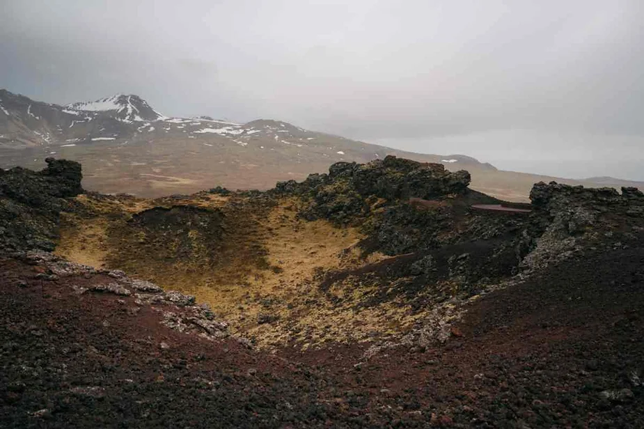 A vast, rugged volcanic landscape in Iceland, featuring a large crater with dark lava rocks and patches of snow-capped mountains in the background under a cloudy sky.