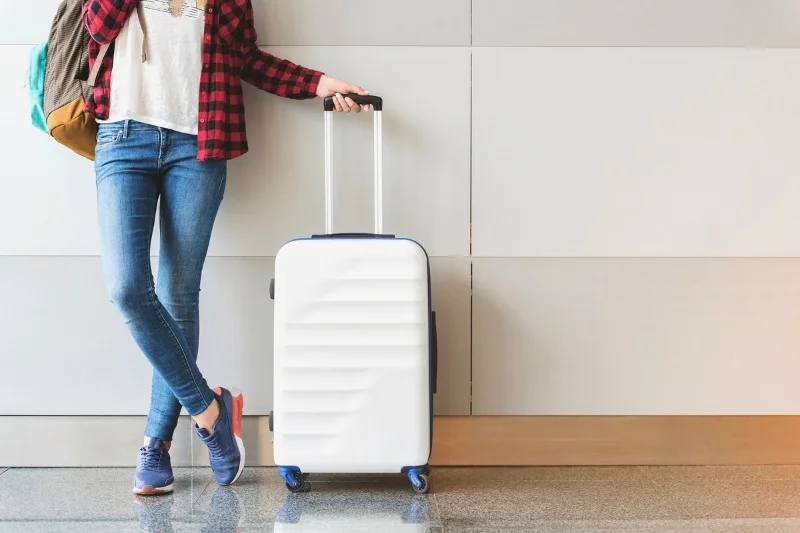 Traveler leaning against a wall, holding the handle of a white suitcase, with a casual outfit of jeans, sneakers, and a plaid shirt.