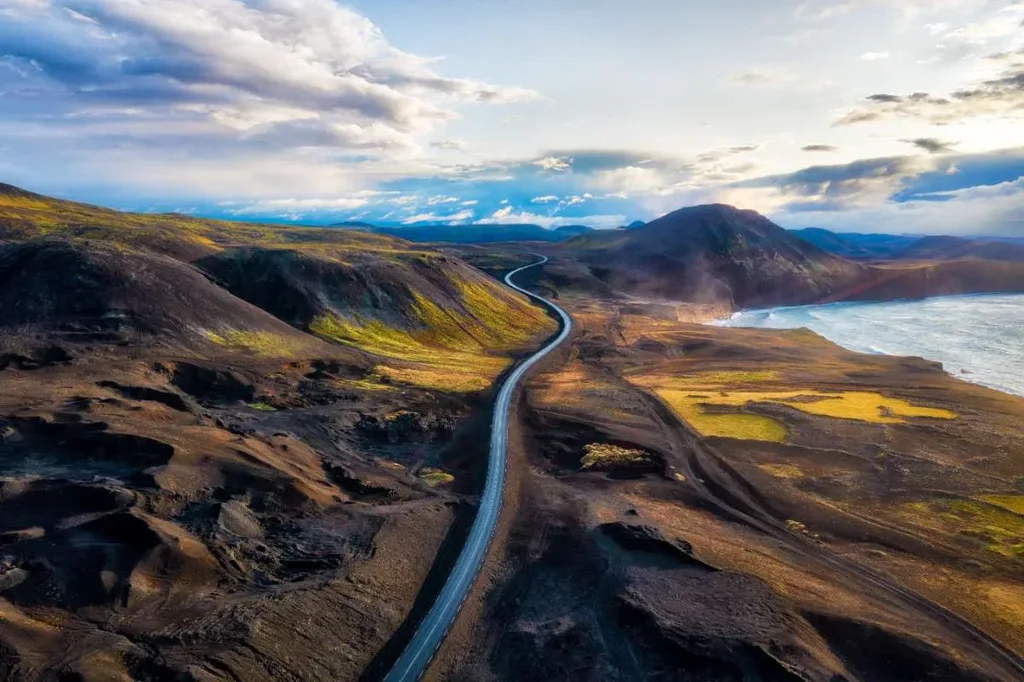 A winding road stretches through the diverse and rugged landscapes of Iceland, with mountains and the ocean in the background under a partly cloudy sky.