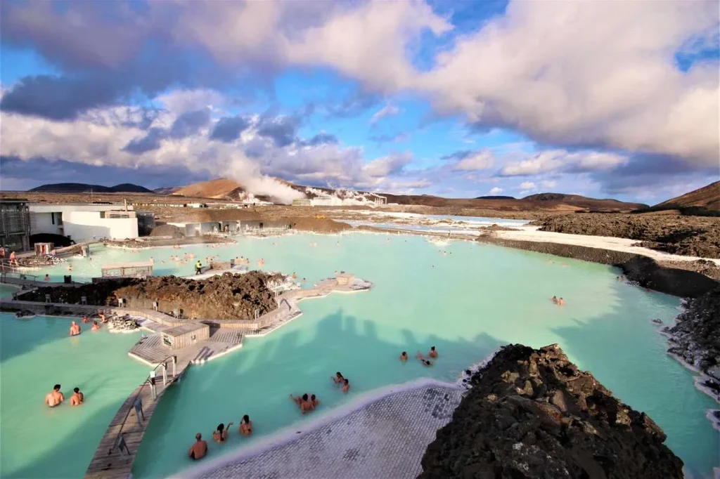 Tourists enjoying a relaxing soak in the Blue Lagoon, a famous geothermal spa in Iceland, surrounded by rocky terrain and set against a backdrop of mountains and a partly cloudy sky.