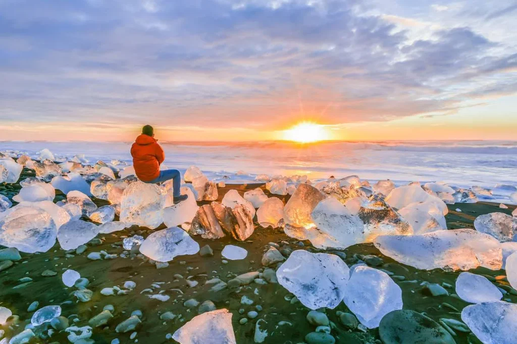 Person in a red jacket sitting on ice formations at Diamond Beach in Iceland, watching a beautiful sunset over the ocean.