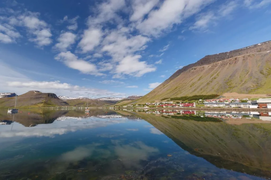 A stunning view of Isafjordur, Iceland, featuring clear blue skies and a tranquil fjord reflecting the surrounding mountains and charming town.