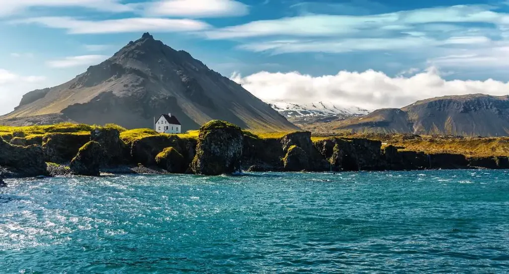 A stunning landscape in Iceland featuring dramatic rock formations along the coastline, with a solitary white house perched atop a hill, framed by mountains and a vibrant blue sky.