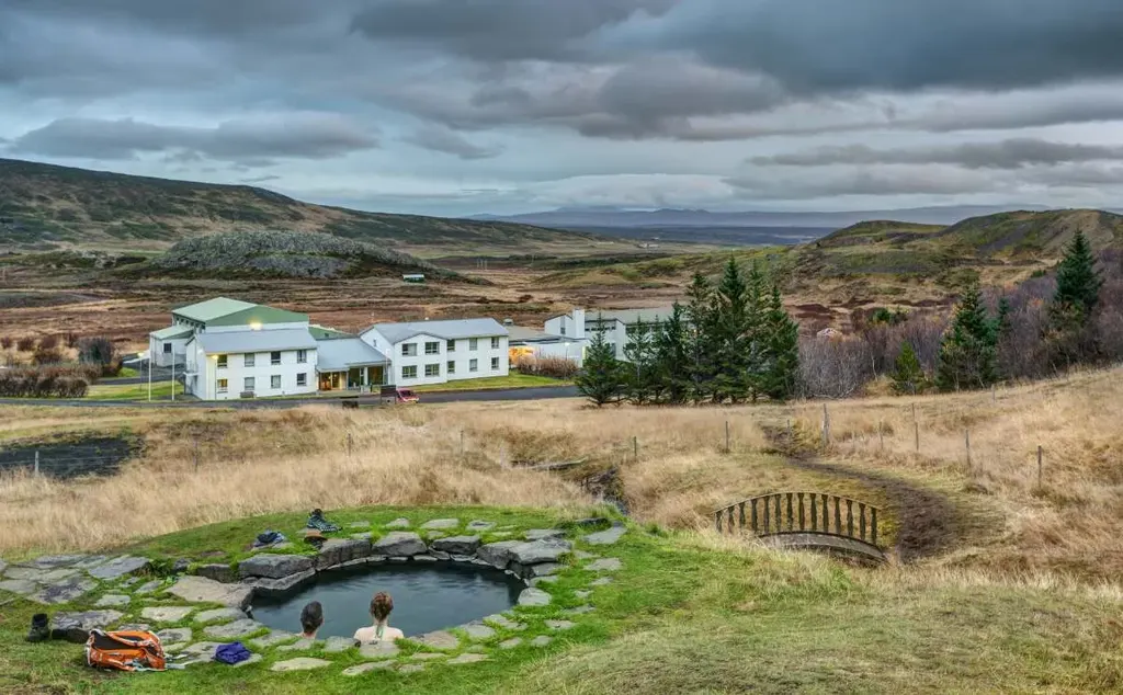 A picturesque view of a thermal bath in Iceland, surrounded by rugged natural landscapes with a few buildings in the background and two people relaxing in the hot spring.