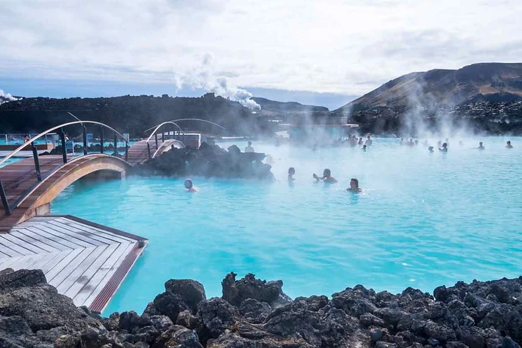 People enjoying the warm waters of a geothermal hot pool in Iceland. The pool is surrounded by rocky terrain with steam rising, and a small bridge crosses over part of the pool.