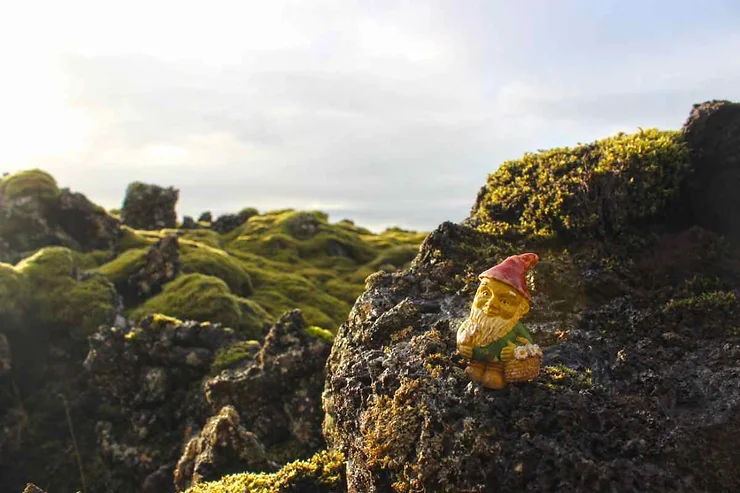 A small elf figurine with a red hat sitting on moss-covered rocks in a scenic Icelandic landscape with a clear sky in the background.