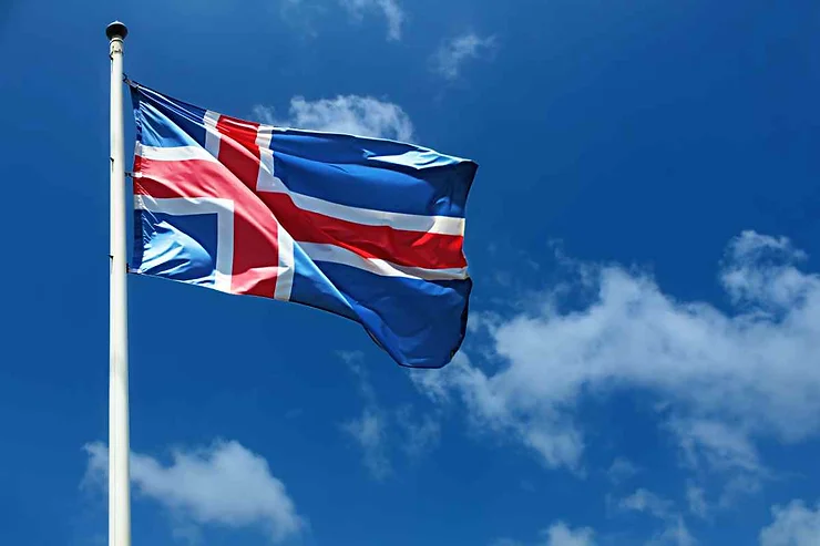 The national flag of Iceland, featuring a blue field with a white and red Nordic cross, waving on a flagpole against a clear blue sky with a few scattered clouds.