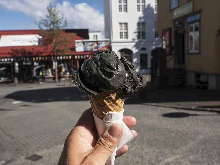 A hand holding a waffle cone with black ice cream in Reykjavik, Iceland. The background shows colorful buildings and a sunny day.