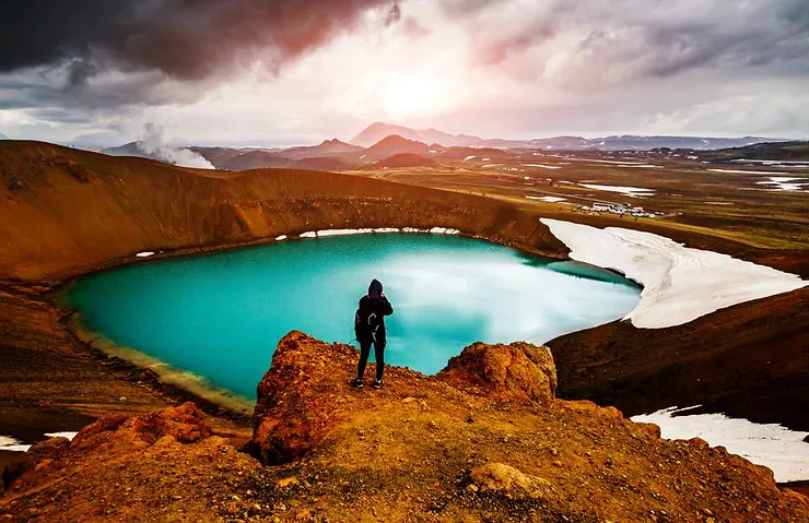 A person stands on a rocky outcrop overlooking a vibrant turquoise volcanic crater lake in Iceland. The landscape around the lake is dramatic, with rugged terrain and distant mountains under a cloudy sky.