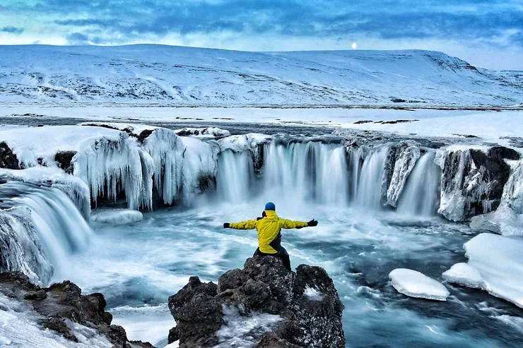 A person in a yellow jacket stands on a rock with arms outstretched, facing a beautiful frozen waterfall in a snowy landscape in Iceland.