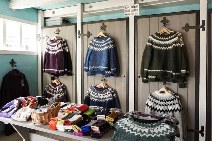 A display of traditional Icelandic woolen sweaters and accessories in a shop. Various sweaters in different colors are hanging on wooden racks, with a table beneath showcasing knitted gloves, hats, and other woolen items.