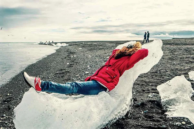 A person in a red jacket lies back on a large chunk of ice on a black sand beach in Iceland. The sky is overcast, and in the distance, other people can be seen walking along the shore.