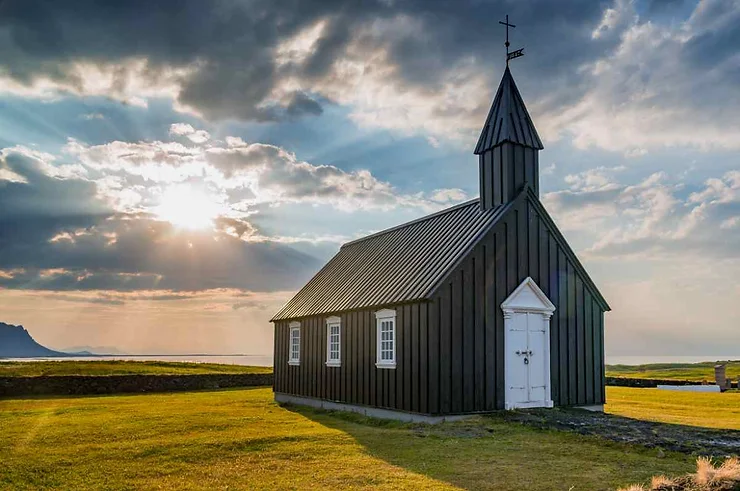 A picturesque view of a small, black wooden church with a white door and windows, set against a backdrop of a stunning sunset with clouds and sun rays breaking through. The church is situated on a grassy field with mountains visible in the distance.