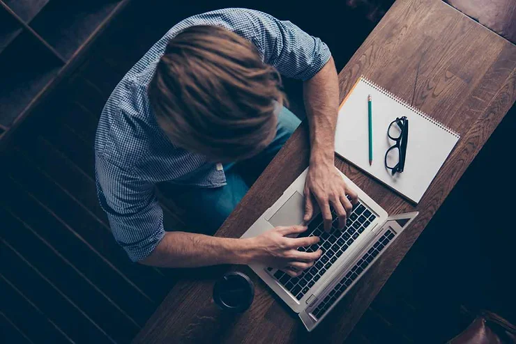 Top view of a man typing on a laptop at a wooden table, with a notebook, glasses, pencil, and a cup of coffee nearby.