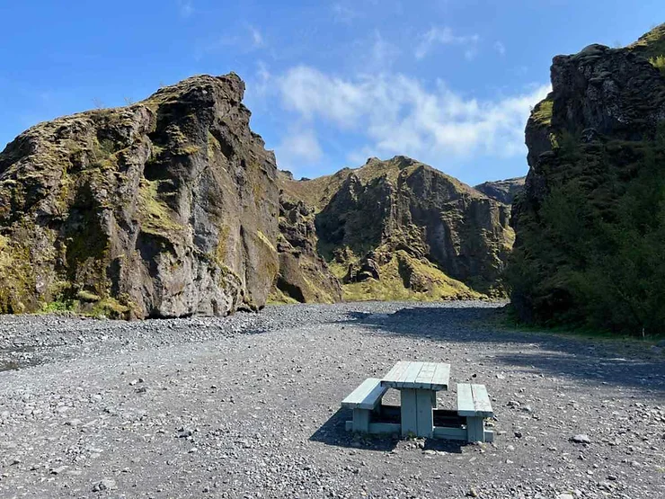 A serene view of Stakkholtsgjá Canyon in Iceland featuring rugged cliffs covered in moss and grass. In the foreground, there is a solitary picnic table on a rocky ground, under a bright blue sky with scattered clouds. The scene evokes a sense of tranquility and natural beauty.