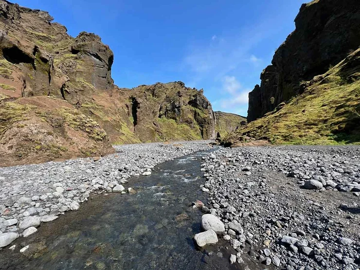 A picturesque view of the Stakkholtsgjá Canyon Trail in Iceland, featuring a narrow stream flowing through a rocky valley surrounded by tall, rugged cliffs covered in moss and grass. The sky is a clear blue, adding to the tranquil and scenic atmosphere of the canyon.