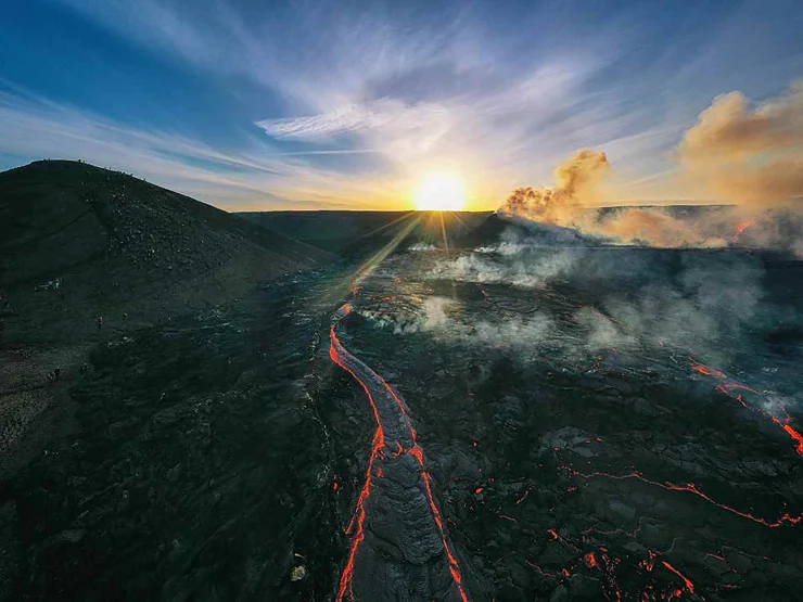 An aerial view of a volcanic eruption in Iceland with flowing lava streams and smoke rising from the ground. The sun is setting in the background, casting a dramatic light over the rugged landscape, emphasizing the raw power and beauty of nature.