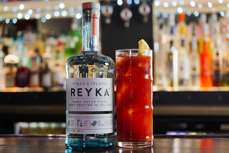 A bottle of Reyka small batch vodka and a cocktail glass with a red drink garnished with a lemon slice, set on a bar counter with blurred bottles and lights in the background in Iceland.